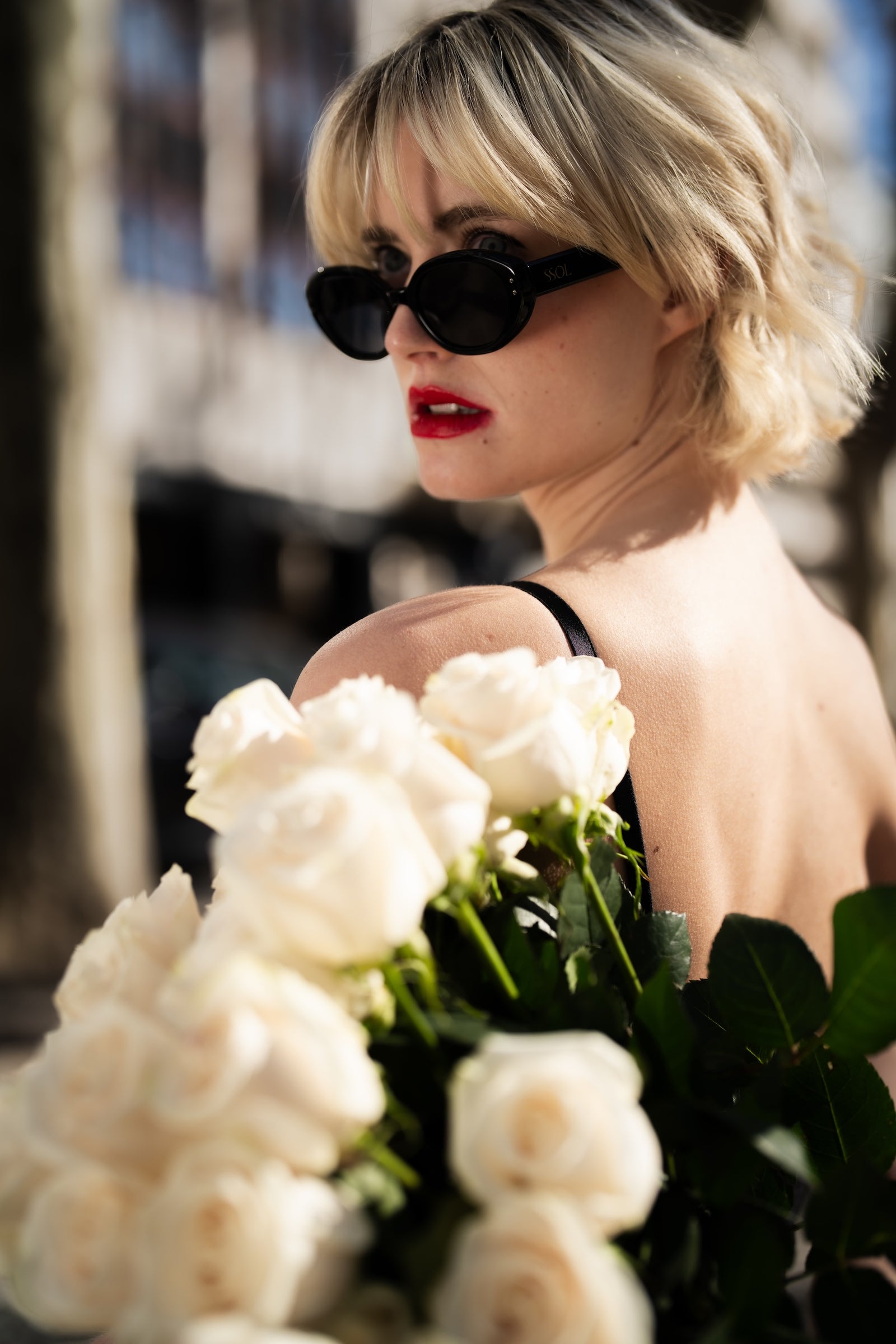 Mulher jovem com cabelo loiro curto usando óculos de sol escuros e batom vermelho, segurando um buquê de rosas brancas. Ela veste um vestido preto com alças finas em um ambiente urbano desfocado ao fundo.
